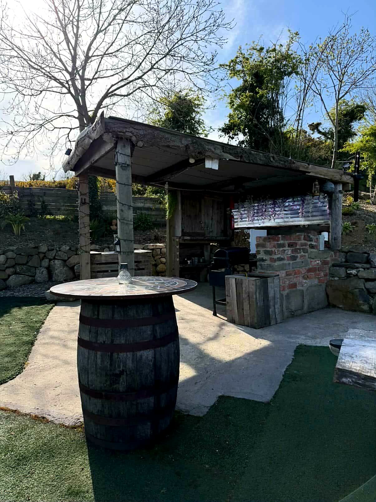 Timber-roofed cooking shelter with barbecue and a whiskey-barrel table beside Fairy Cove
