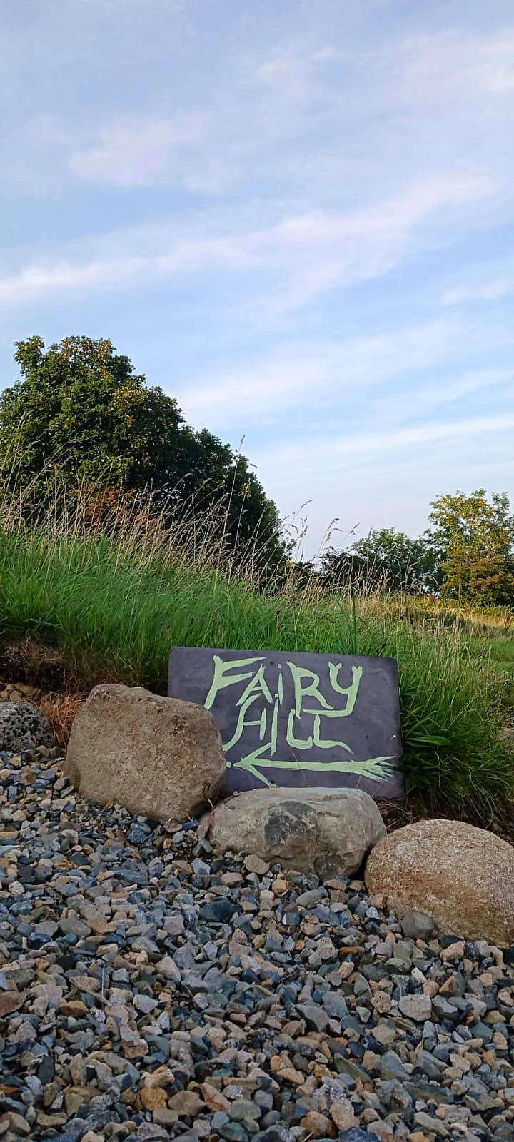 Hand-painted Fairy Hill slate sign set among stones and gravel, with long grass and trees behind