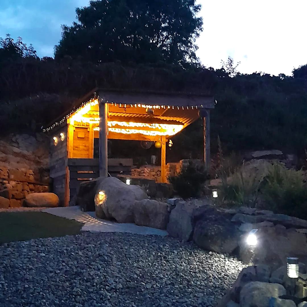 Fairy Hill's timber cooking shelter glowing with fairy lights at dusk among stone walls and planting